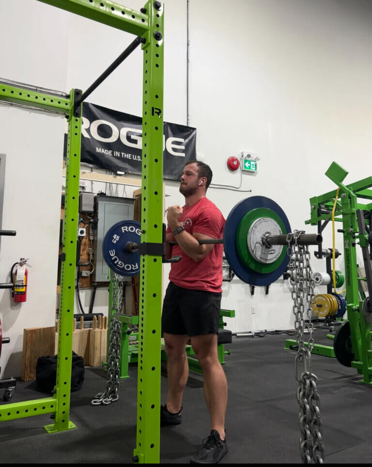 Man in red shirt preparing for a Zercher squat, gripping a heavy barbell with chains hanging, showcasing strength and determination.